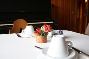 Una mesa dispuesta con tazas blancas, un cuenco de madera con flores y un piano al fondo.