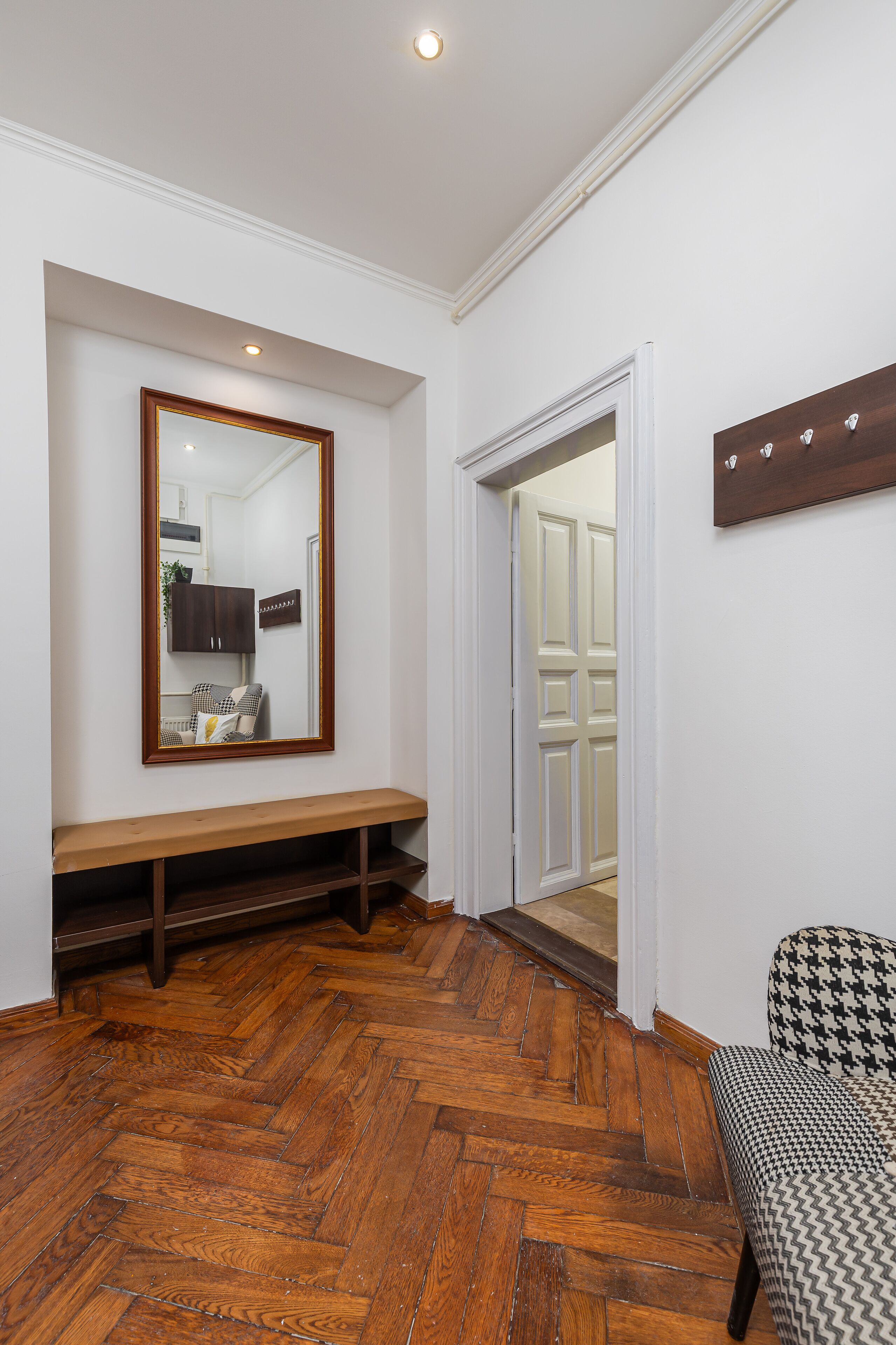 Un couloir doté d’un parquet en bois disposé en point de Hongrie, d’un banc en bois, d’un grand miroir et d’une porte donnant sur une autre pièce.