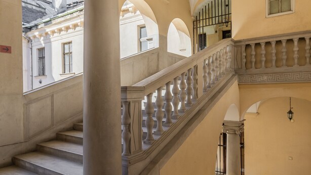 Una imponente escalera con columnas clásicas y barandilla, que conduce a un piso superior en un edificio con ventanas arqueadas y fachada amarilla.