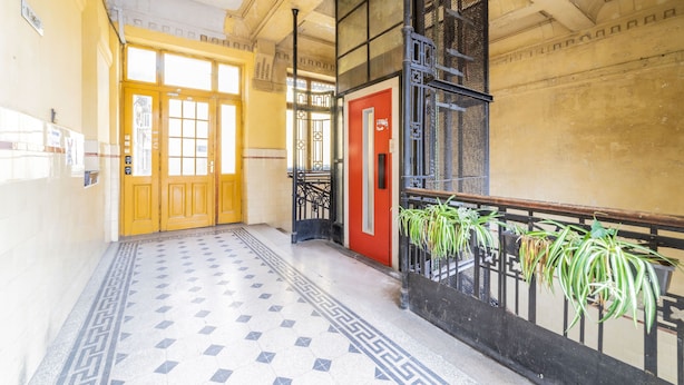 A hallway with patterned floor tiles, a red door, and a black railing with potted plants. - Real Apartments Anker koz (Budapest)