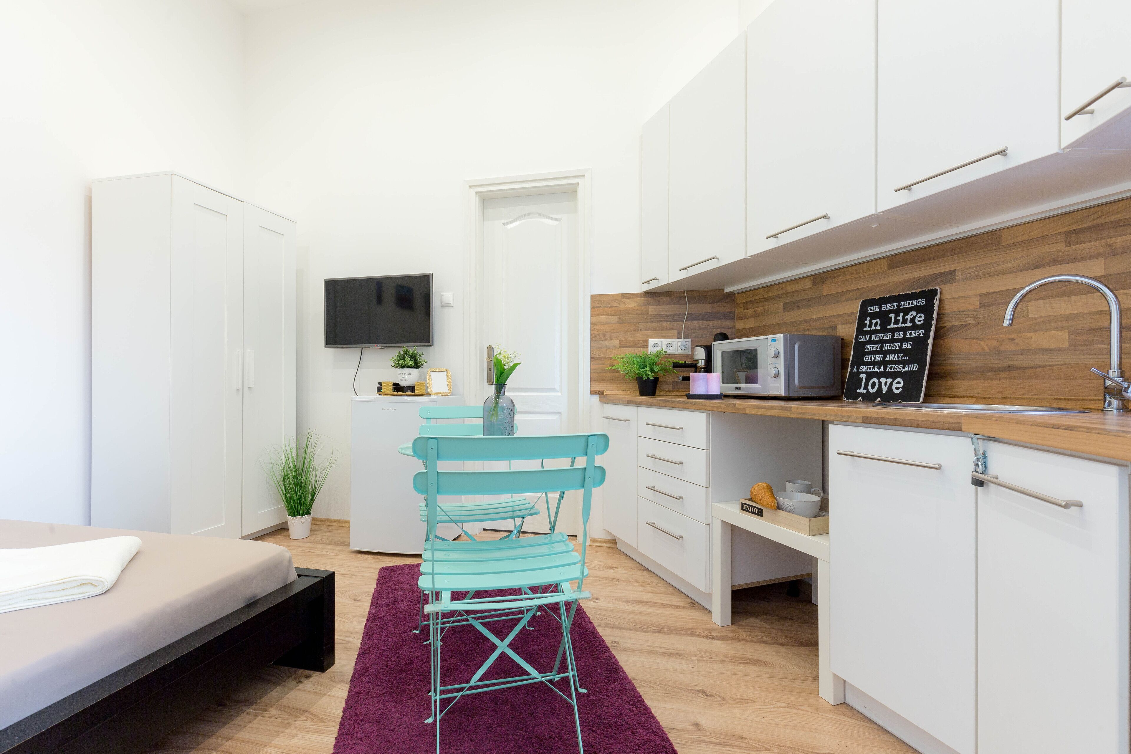 Habitación de hotel compacta con cama, kitchenette, televisor y pequeña zona de comedor con una silla y mesa.