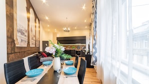 A dining area with a long table set for four, a modern kitchen in the background, and a large window with sheer curtains. - Real Apartments Anker koz (Budapest)