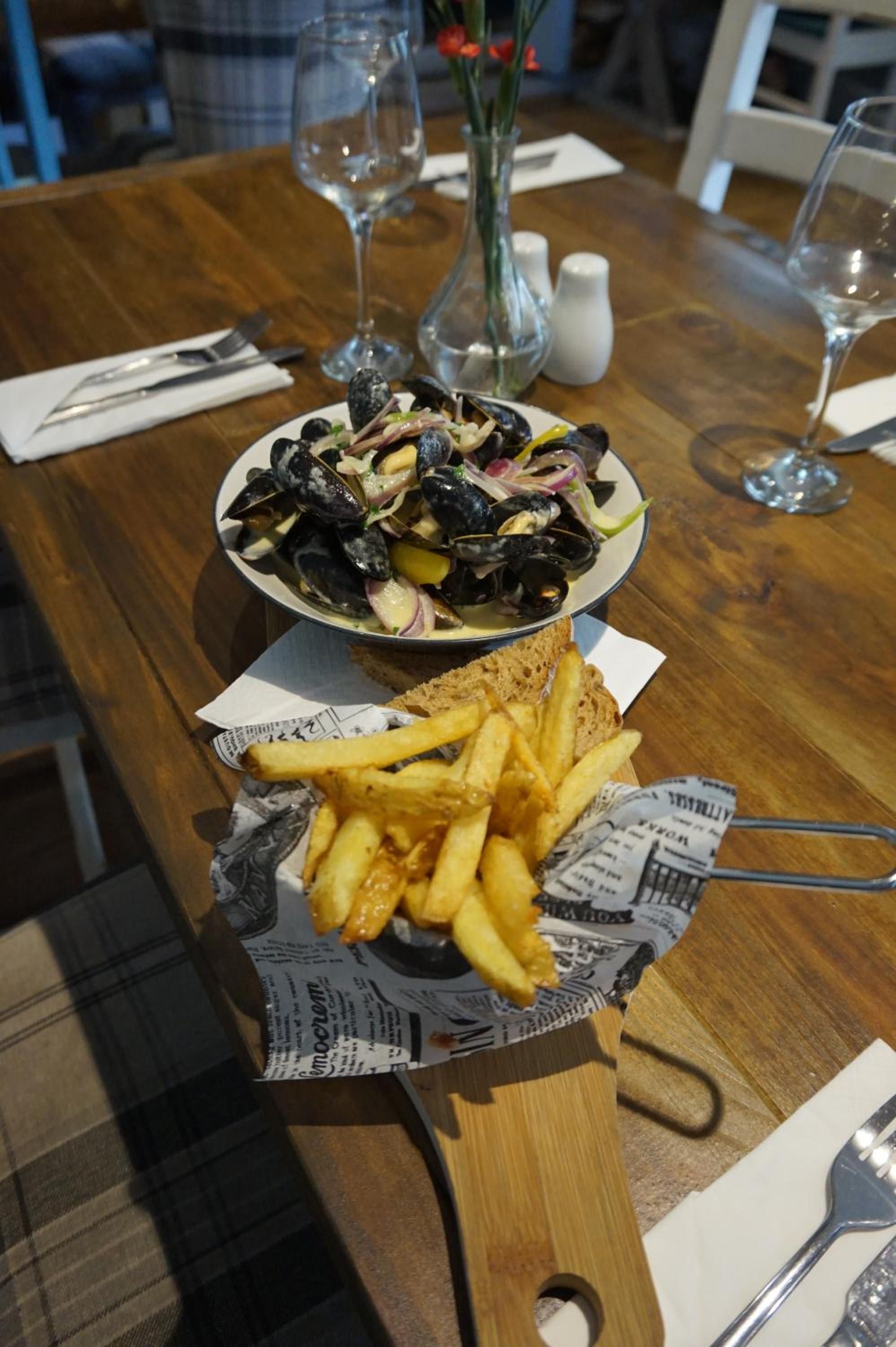 A dining table with a plate of mussels, a basket of fries, and a newspaper.