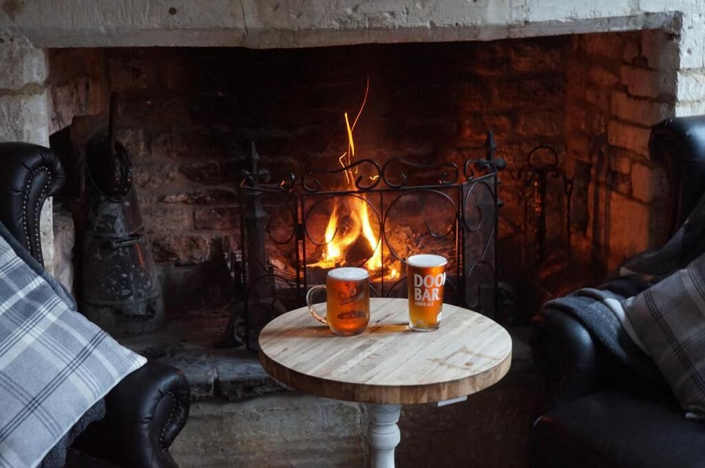 A fireplace with two mugs on a round table, a chair with a plaid cushion, and a brick wall.