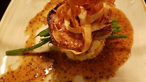 Un plato con un guiso que incluye un trozo de carne, posiblemente cerdo, cubierto de cebollas fritas crujientes y adornado con hierbas verdes.