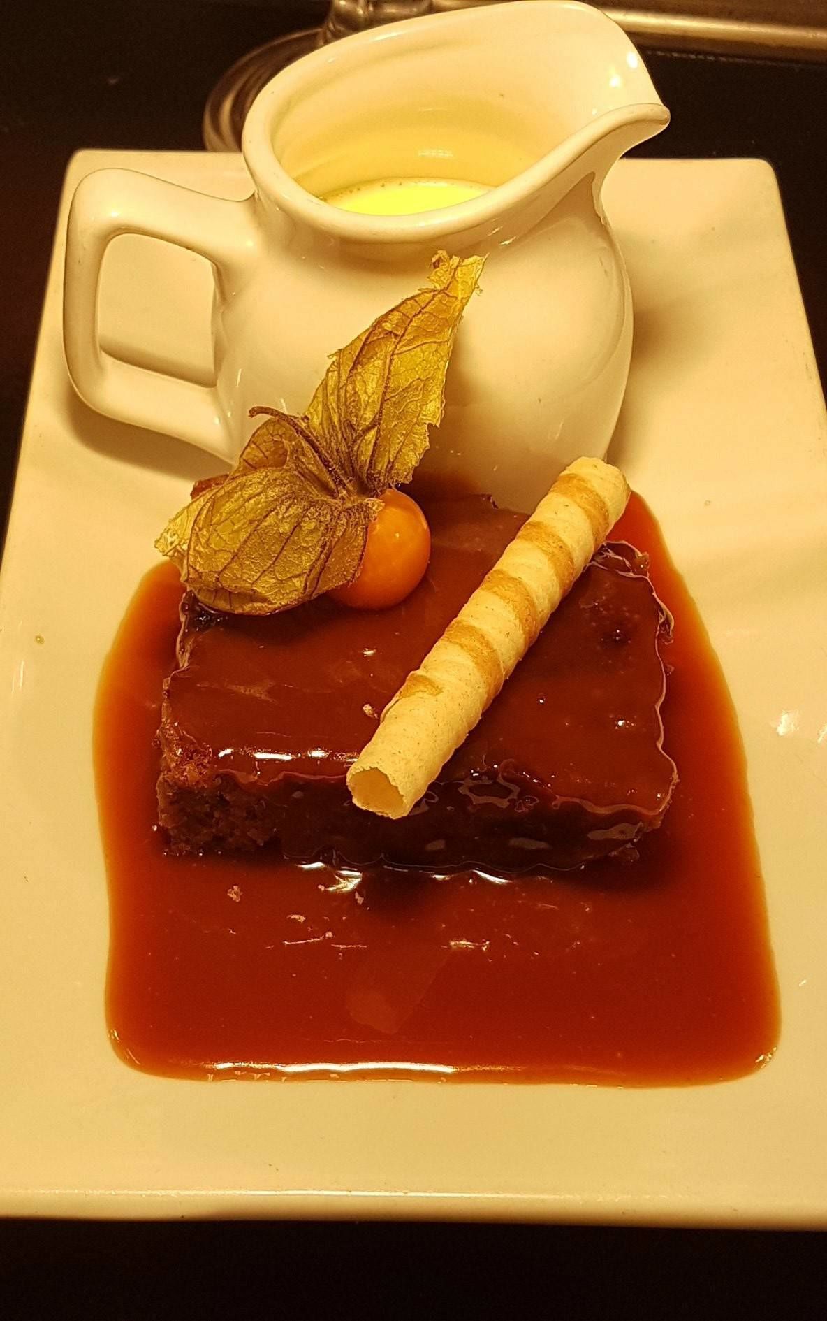 A dessert plate with a slice of cake, a small pitcher of sauce, and a decorative leaf.