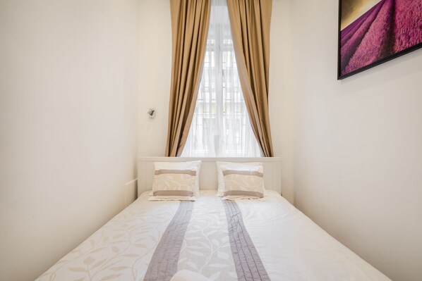 Un dormitorio con cama, dos almohadas, ventana con cortinas y un cuadro en la pared.