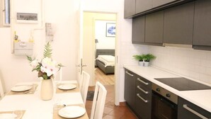 Une cuisine compacte avec des meubles sombres, un plan de travail et une cuisinière. On y trouve un coin repas avec une table dressée pour deux personnes, un vase avec des fleurs et une porte donnant sur une chambre.