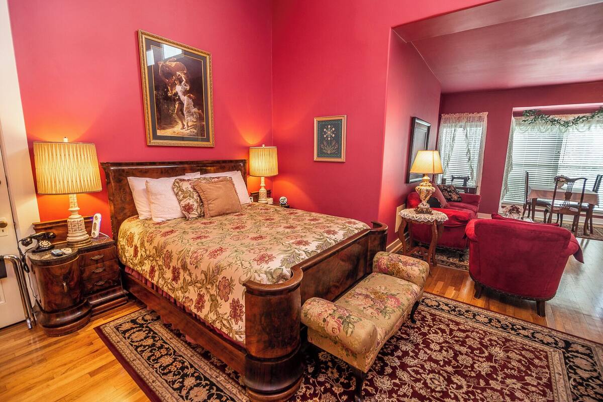 Une chambre avec un grand lit, deux tables de chevet avec des lampes, un fauteuil rouge, un banc et un coin repas avec une table et des chaises.