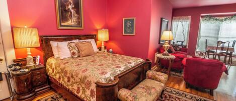 A bedroom with a large bed, two bedside tables with lamps, a red armchair, a bench, and a dining area with a table and chairs.