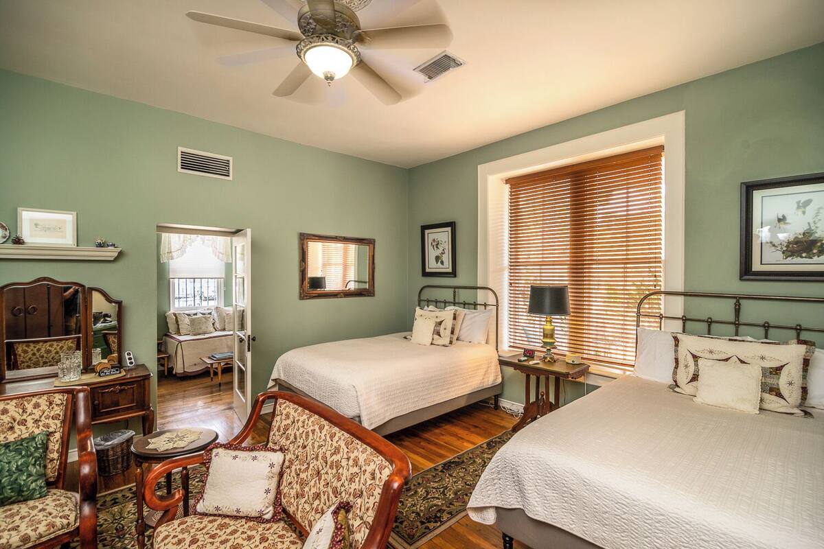 Une chambre avec deux lits, un ventilateur de plafond, une fenêtre avec des stores, une table de chevet avec une lampe et un miroir au mur.