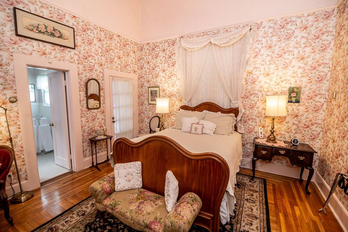 Une chambre avec un papier peint à motifs floraux, un lit en bois, une table de chevet, une chaise et un miroir.