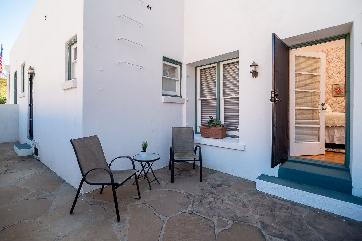 A white building with a stone patio, two chairs, a small table, and a door leading to a bedroom with a floral wallpaper.