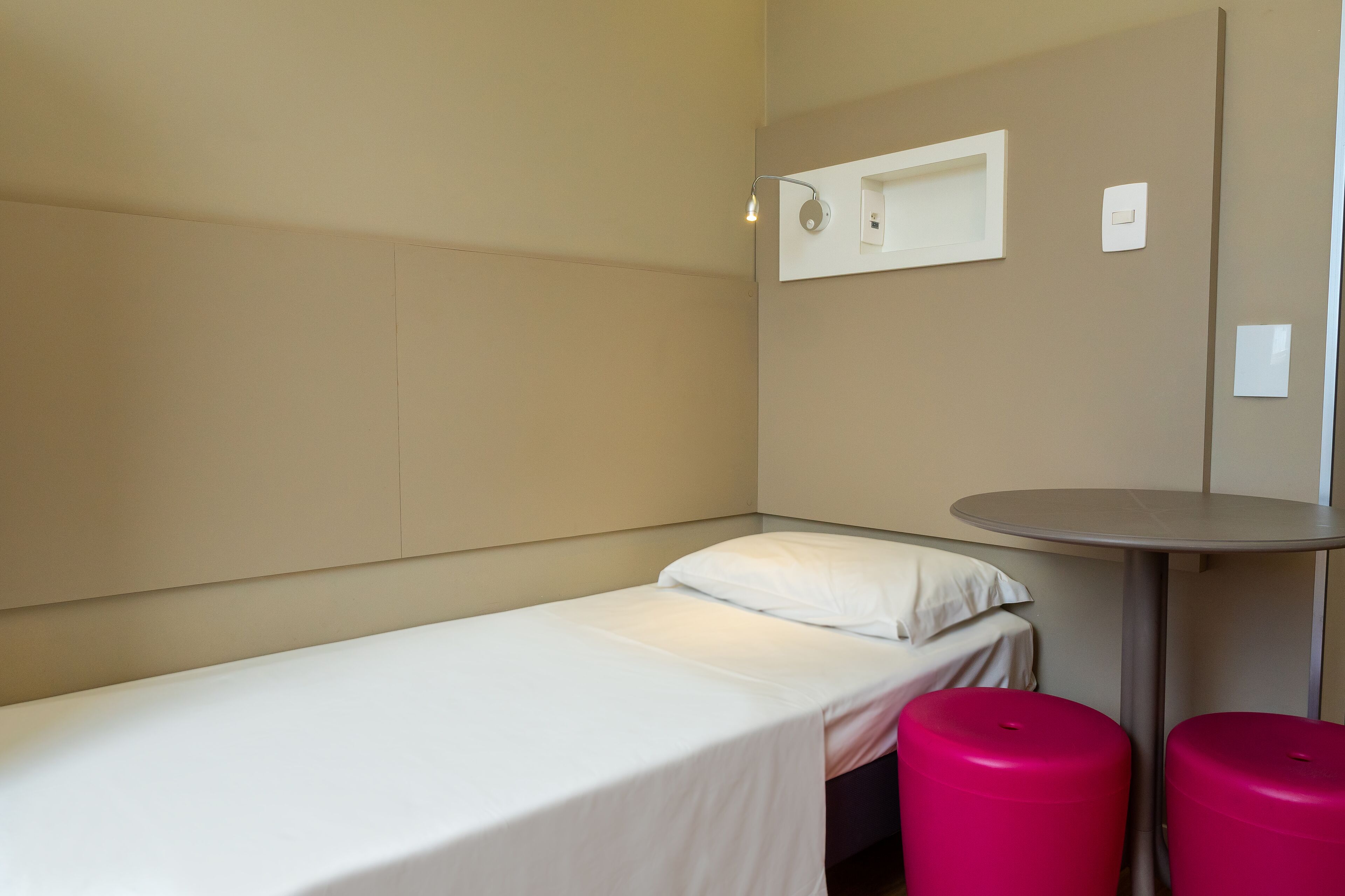 A single bed with a white mattress and pillow, a small table with a round top, and two pink stools.