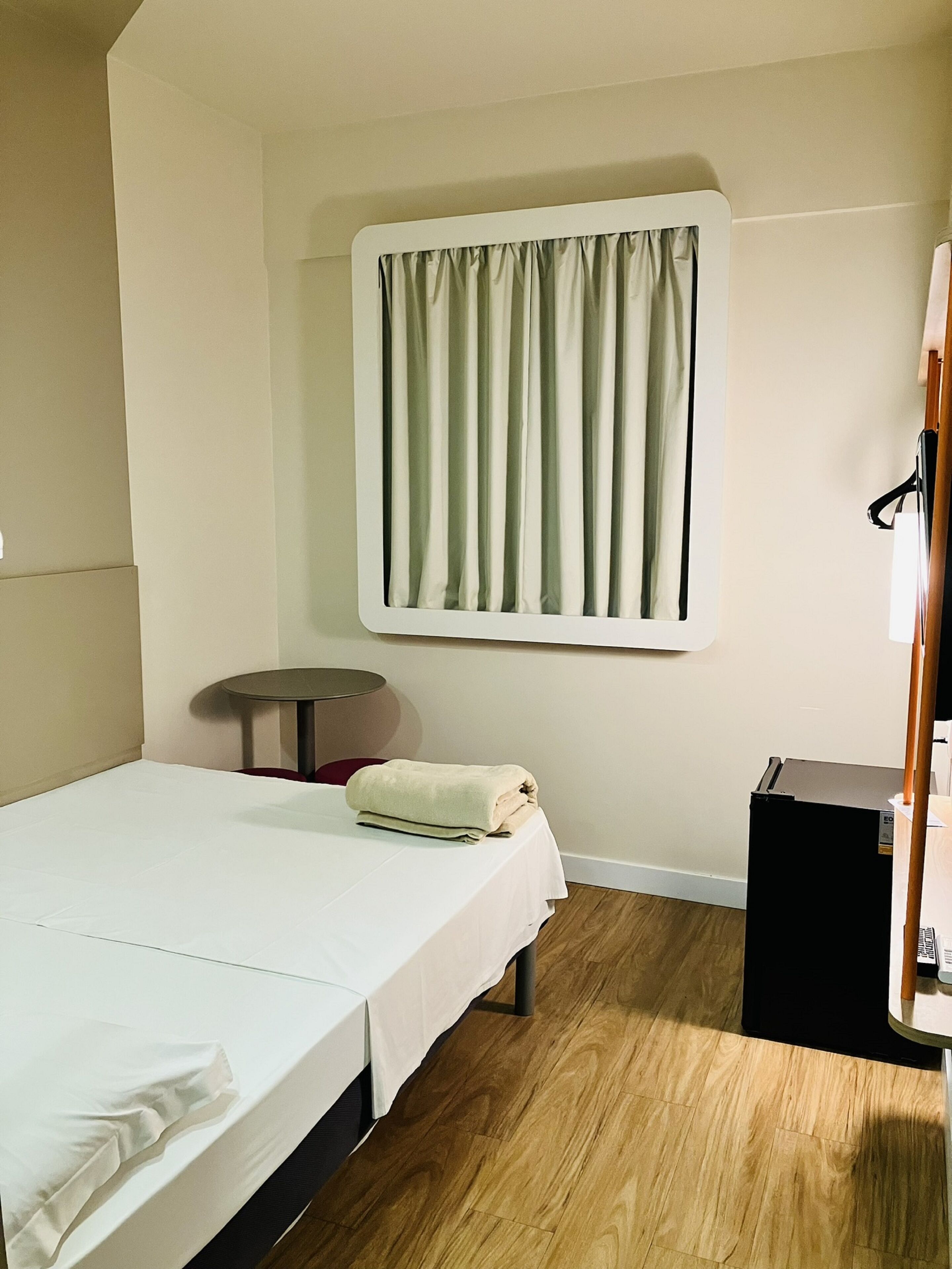 A small, neatly arranged room with a bed, a small table, a window with curtains, and a compact refrigerator.