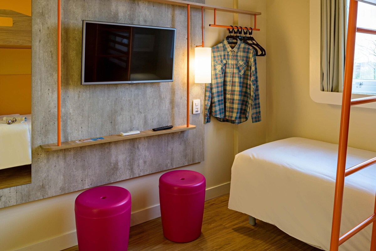 Een kleine kamer met een bed, een televisie bevestigd aan een betonnen muur, een houten plankje met een afstandsbediening, en een kledingrek met een geruite blouse.