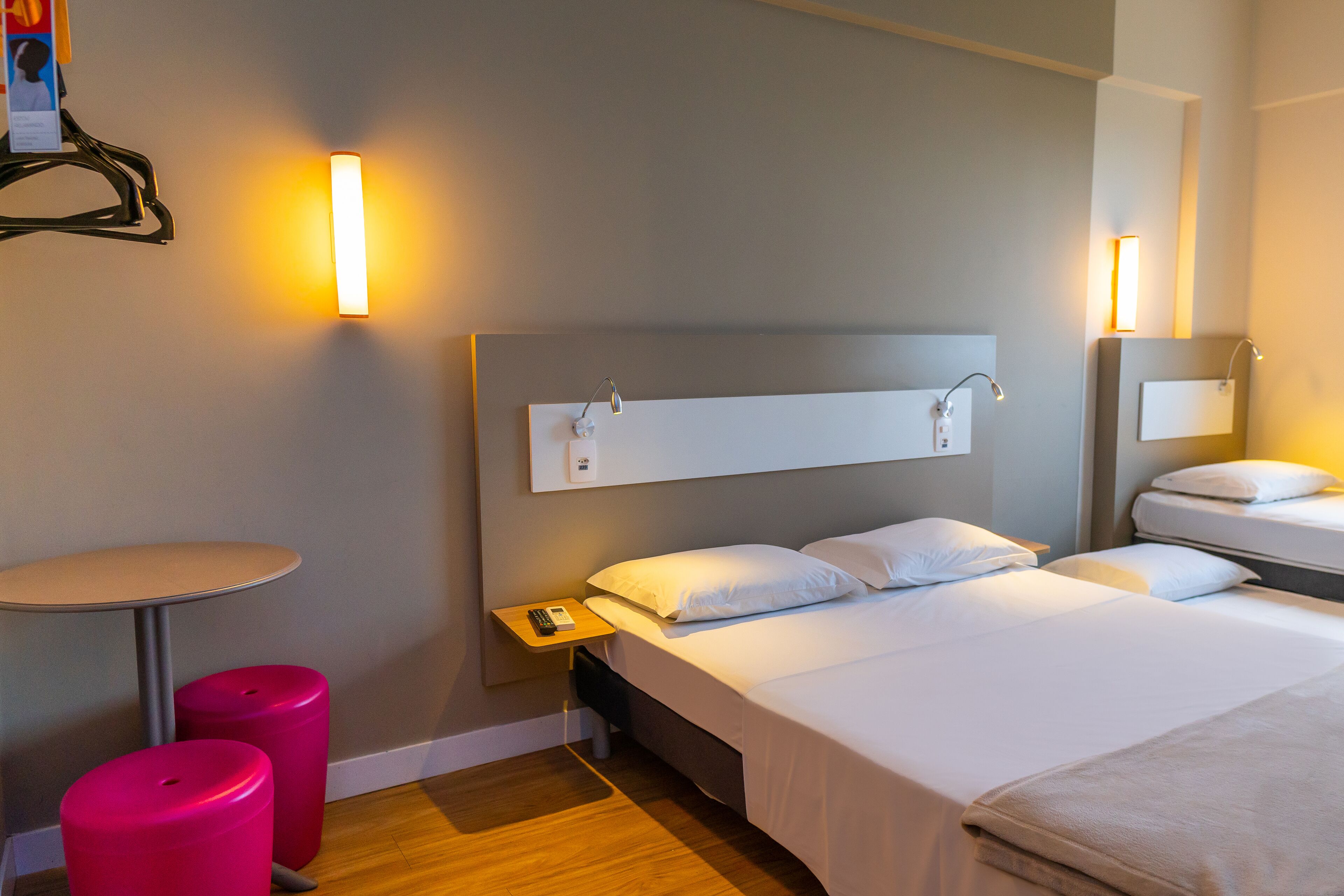 A hotel room with a bed, bedside tables, a small table, and two pink stools.