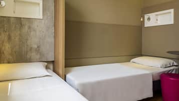 Quarto de hotel com duas camas de solteiro, cabeceira de madeira, uma mesinha e um banquinho rosa.