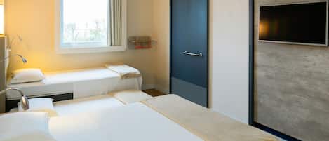 A hotel room with two beds, a window with curtains, a bedside table with a lamp, and a TV mounted on the wall.