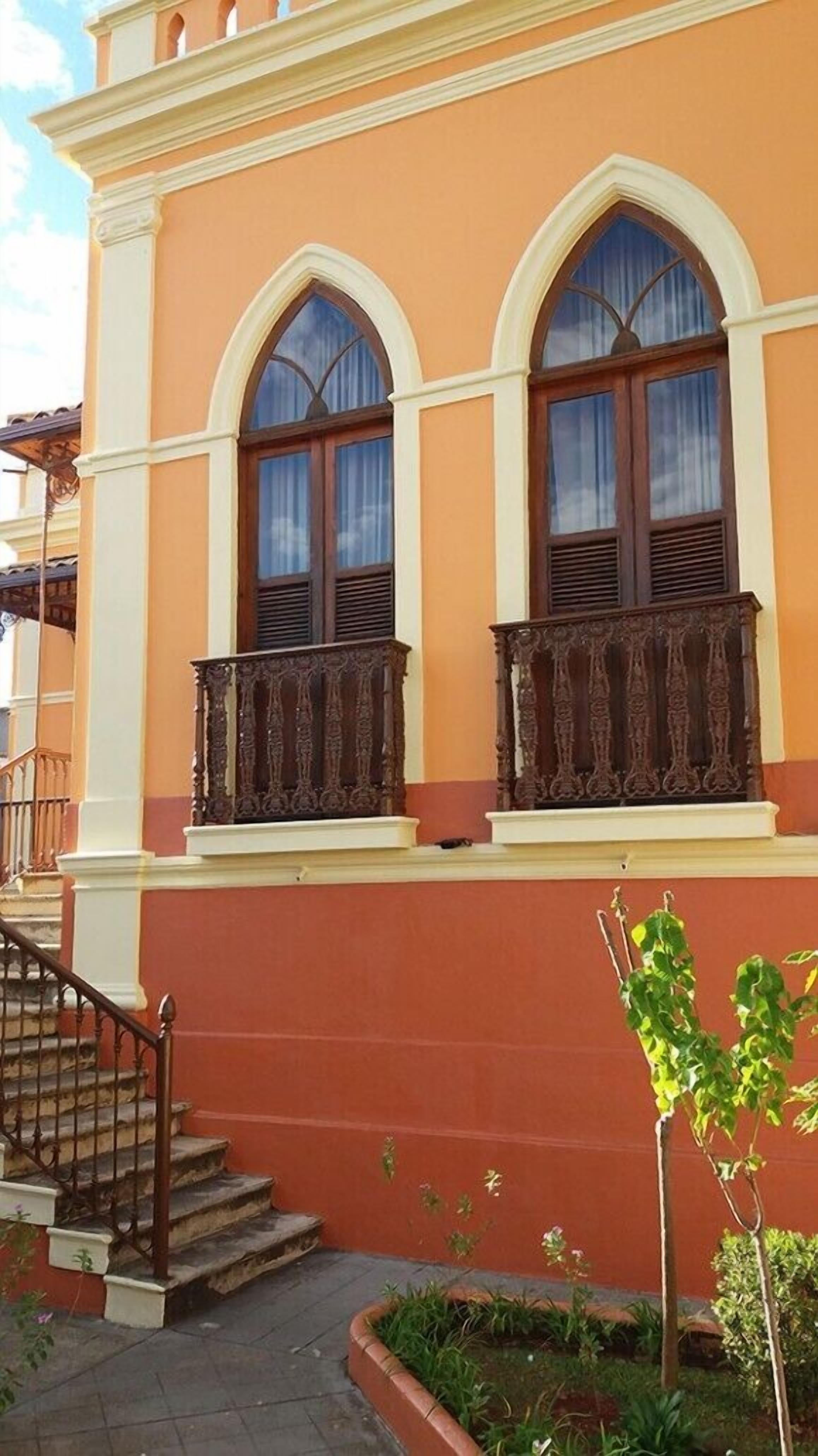 A building with two arched windows, a balcony with ornate railings, and a staircase leading to the entrance.