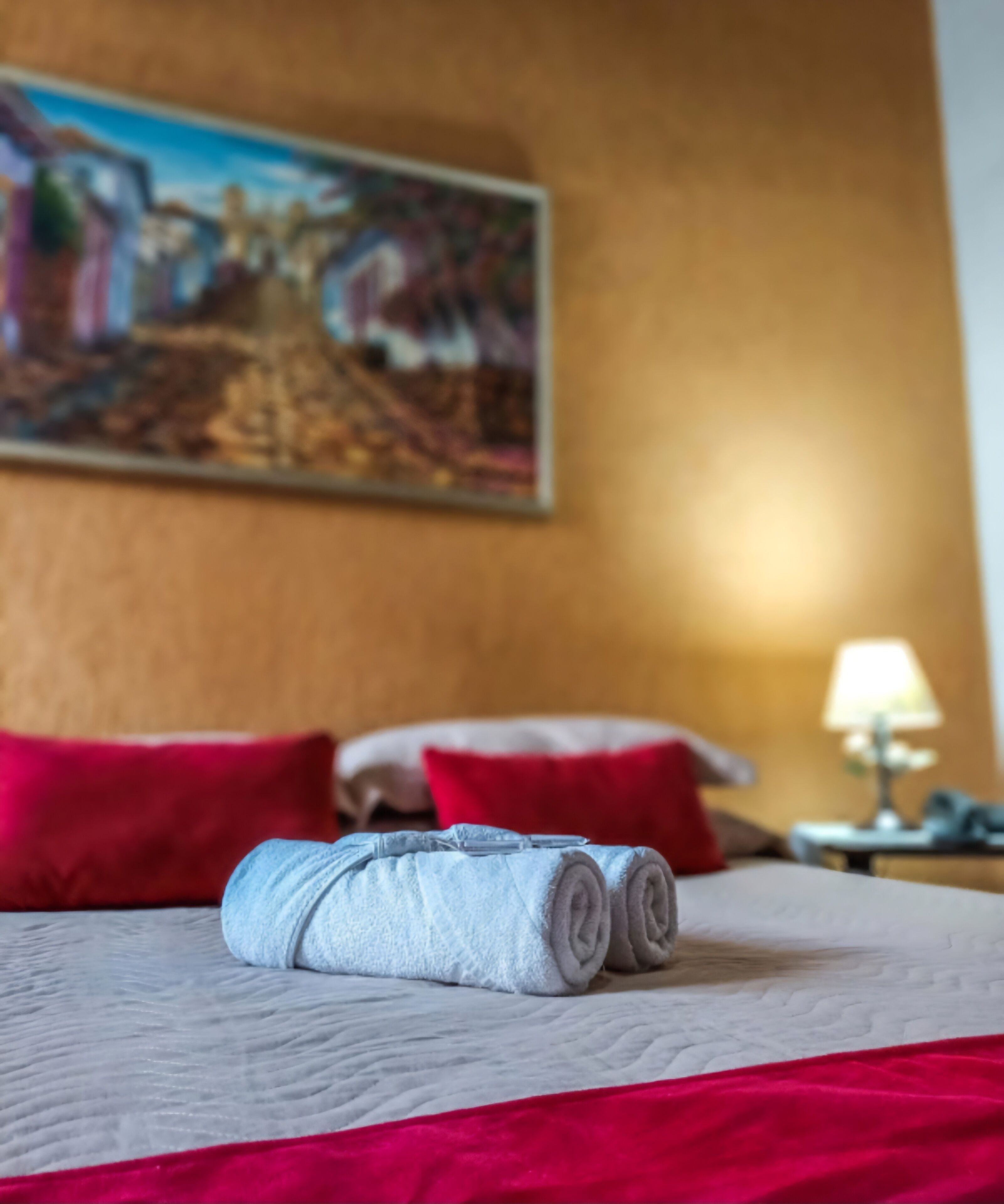 Una cama bien tendida con una toalla enrollada, almohadas rojas y un cuadro en la pared.