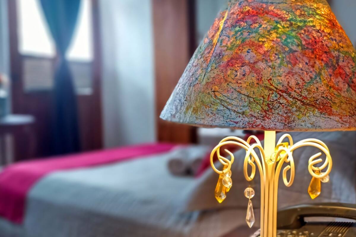 Un lit avec une couverture rose, une lampe avec un abat-jour coloré et une table de chevet en bois.