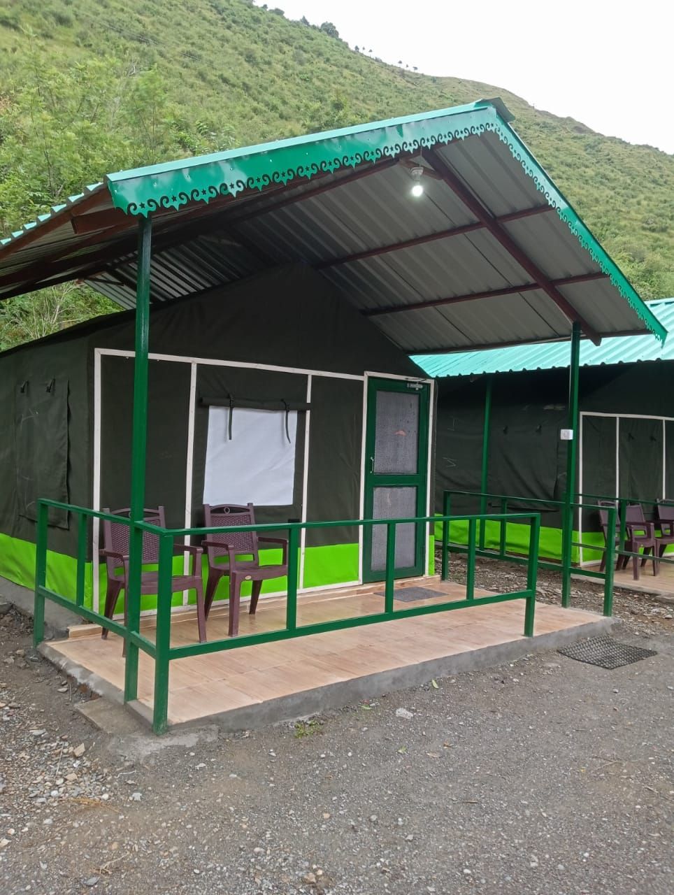 A small, single-story building with a green exterior and a corrugated metal roof. It has a covered outdoor seating area with plastic chairs and a table.