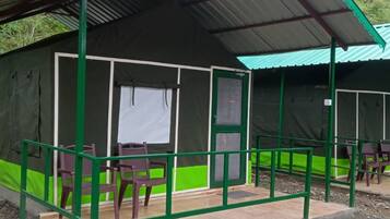 A small, single-story building with a green exterior and a corrugated metal roof. It has a covered outdoor seating area with plastic chairs and a table.