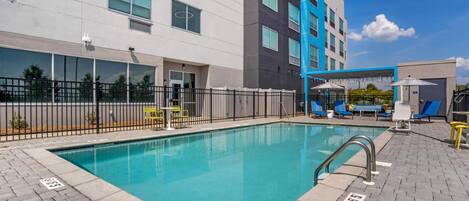A swimming pool with a metal ladder, surrounded by a paved deck with lounge chairs and a fence.