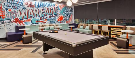 Una mesa de billar en un área de lounge con un mural que presenta temas deportivos y de entretenimiento.