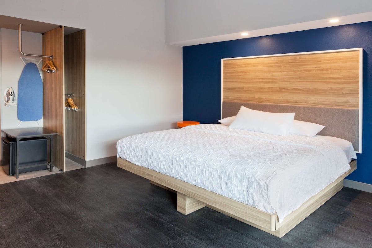 A modern hotel room with a large bed, a wooden headboard, a built-in ironing station, and a dark wooden floor.