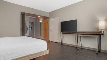 A modern hotel room with a bed, a flat-screen TV, a desk with a lamp, and a shower area.