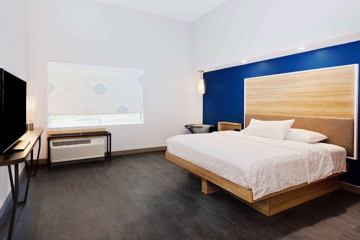 A modern hotel room with a bed, a TV, a heater, and a window with blinds.