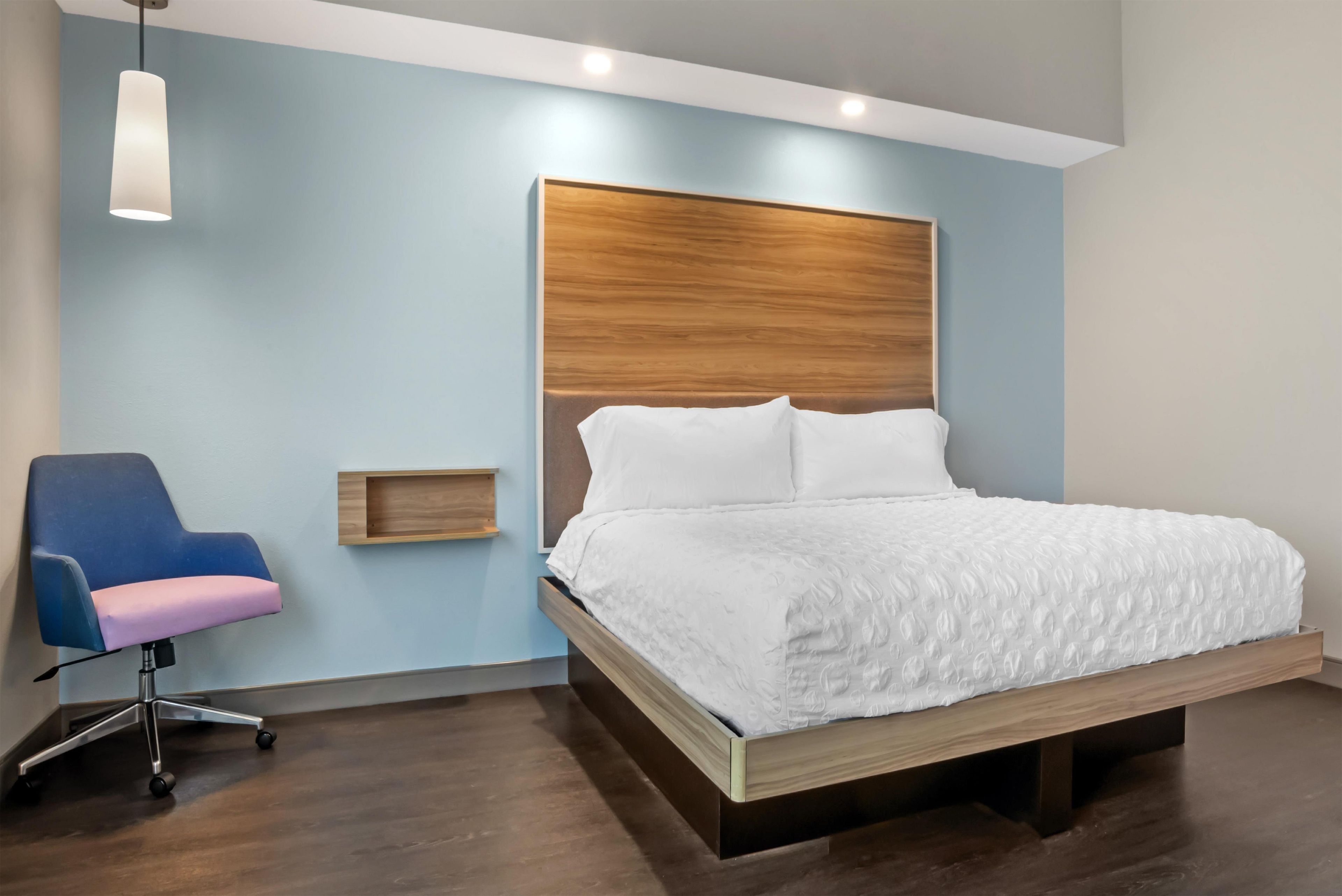 A modern bedroom with a wooden headboard, a single bed with white bedding, a blue office chair, and a pendant light.