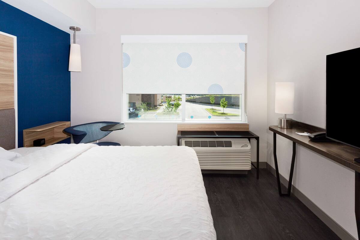 A hotel room with a bed, a desk, a chair, a TV, a window with a view of a parking lot, and a heater.