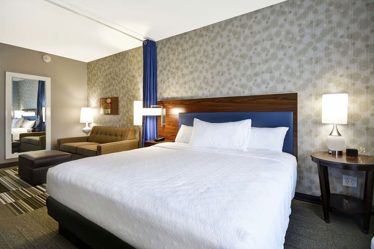 A hotel room with a large bed, a sofa, a chair, a nightstand, a lamp, and a mirror.
