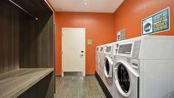 A laundry room with a row of front-loading washers and dryers, a door, and a sign instructing to notify the front desk if stepping out.