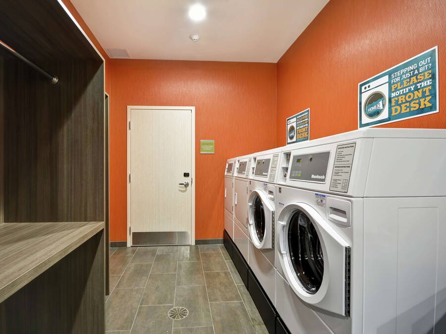 A laundry room with a row of front-loading washers and dryers, a door, and a sign instructing to notify the front desk if stepping out.