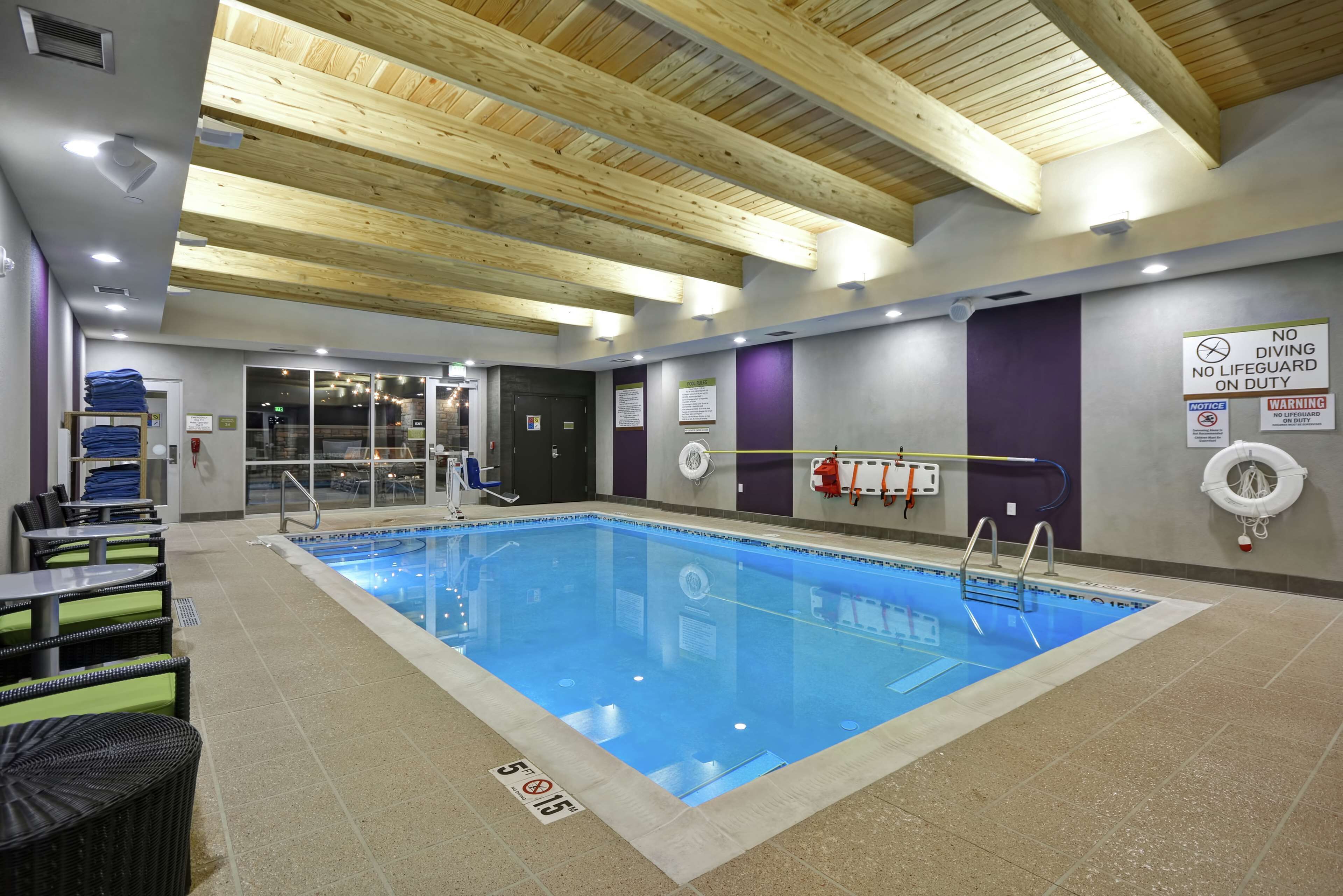 An indoor swimming pool with a wooden ceiling, safety signs, and lounge area with chairs and tables.
