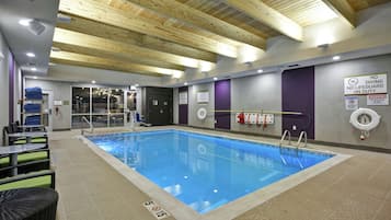 An indoor swimming pool with a wooden ceiling, safety signs, and lounge area with chairs and tables.