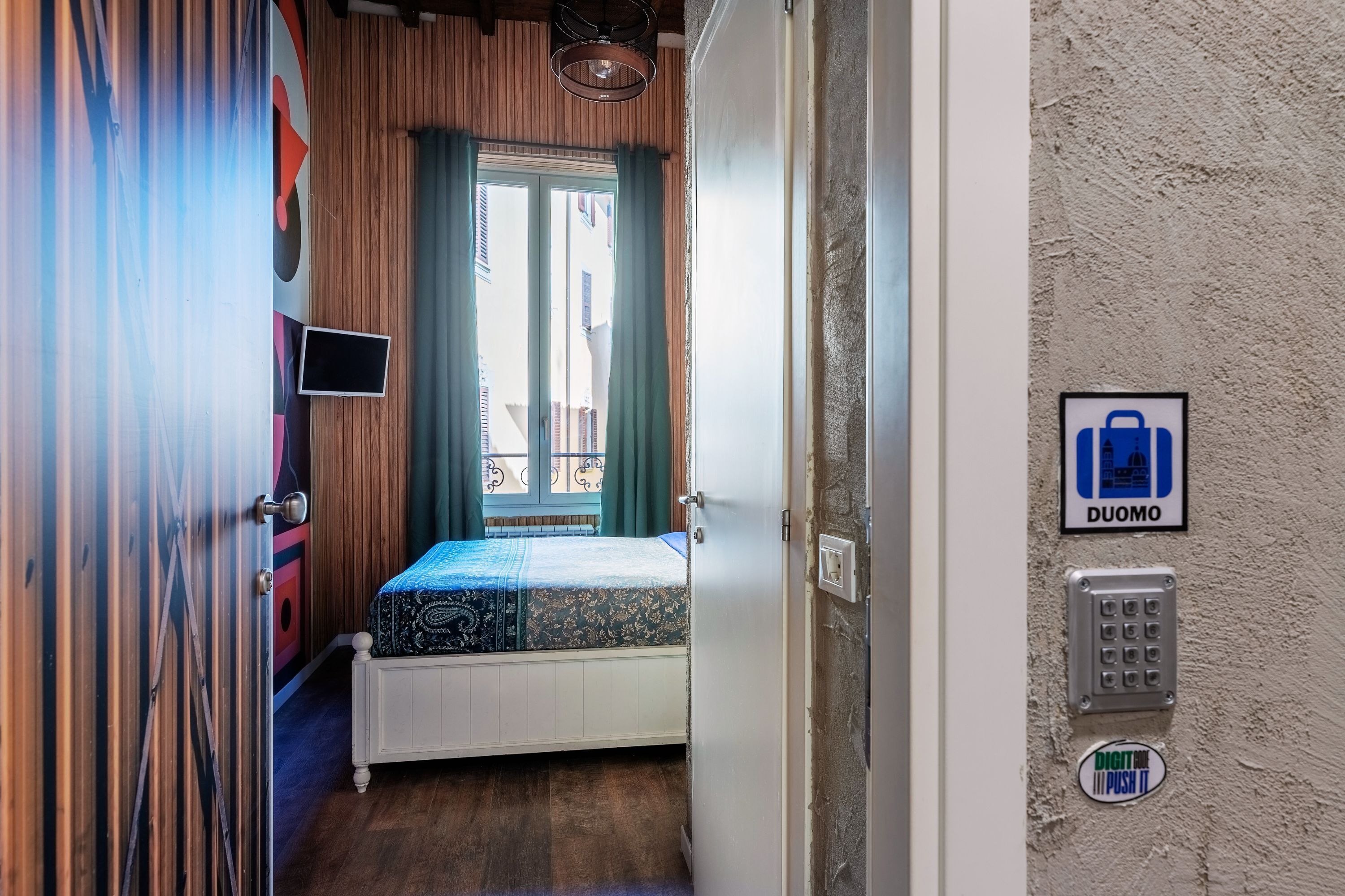 A bedroom with a bed, a window with curtains, a wall-mounted TV, and a door with a handle.