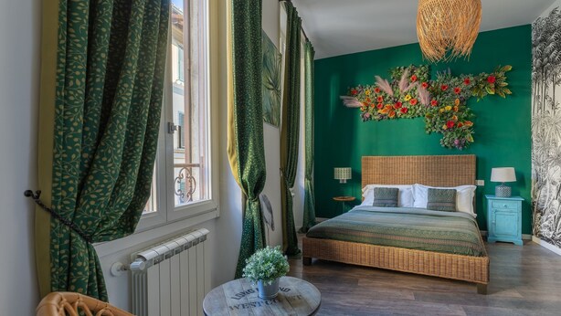 Une chambre avec un mur vert, un lit en osier, une table ronde avec une plante et une fenêtre avec des rideaux.