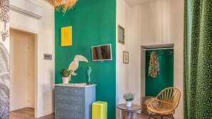 Une pièce avec un mur d’accent vert, une commode grise, une valise jaune, une chaise en osier et une petite table ronde agrémentée d’une plante.