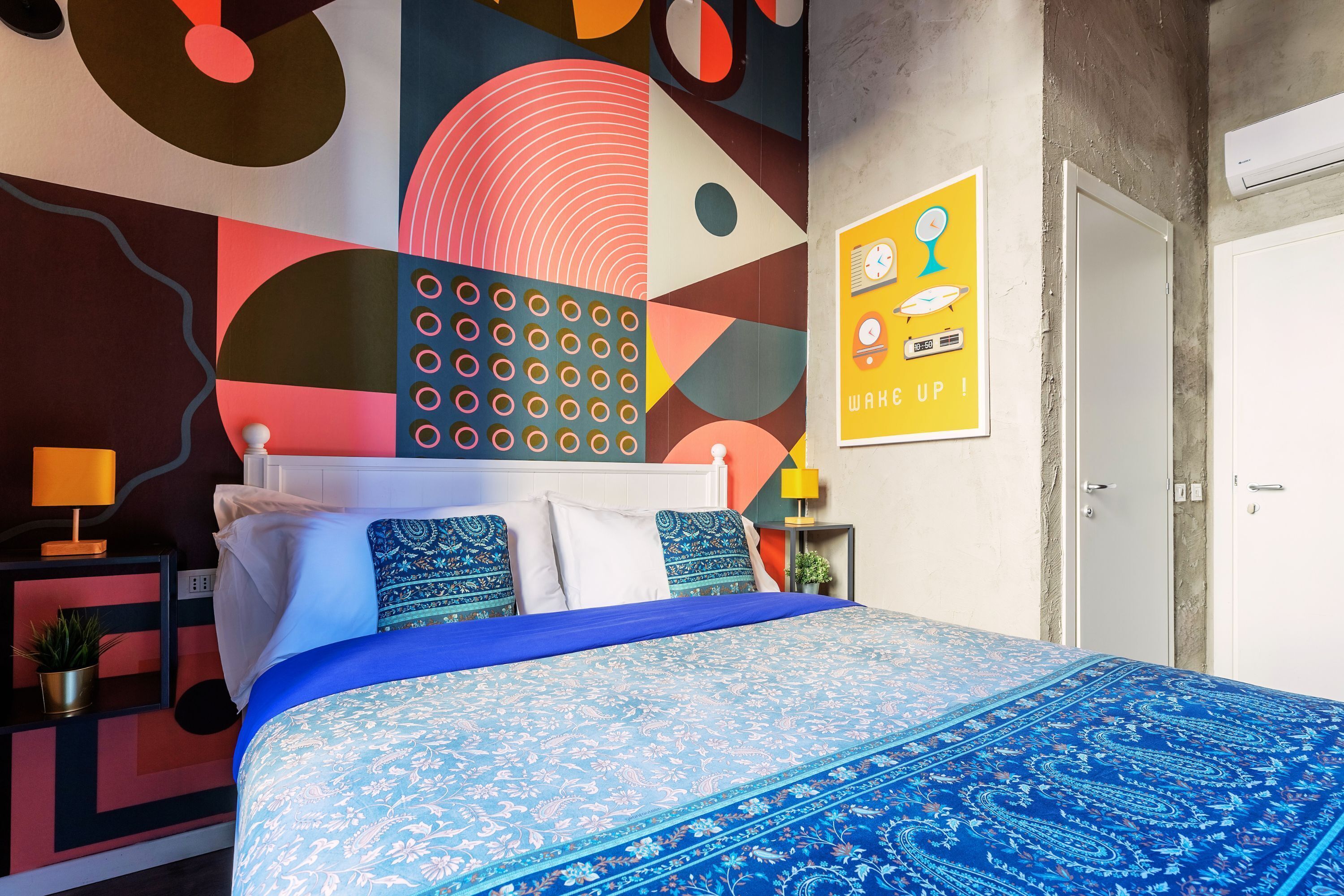 A modern bedroom with a large bed, a colorful abstract wall mural, a clock poster, and a small bedside table with a lamp.