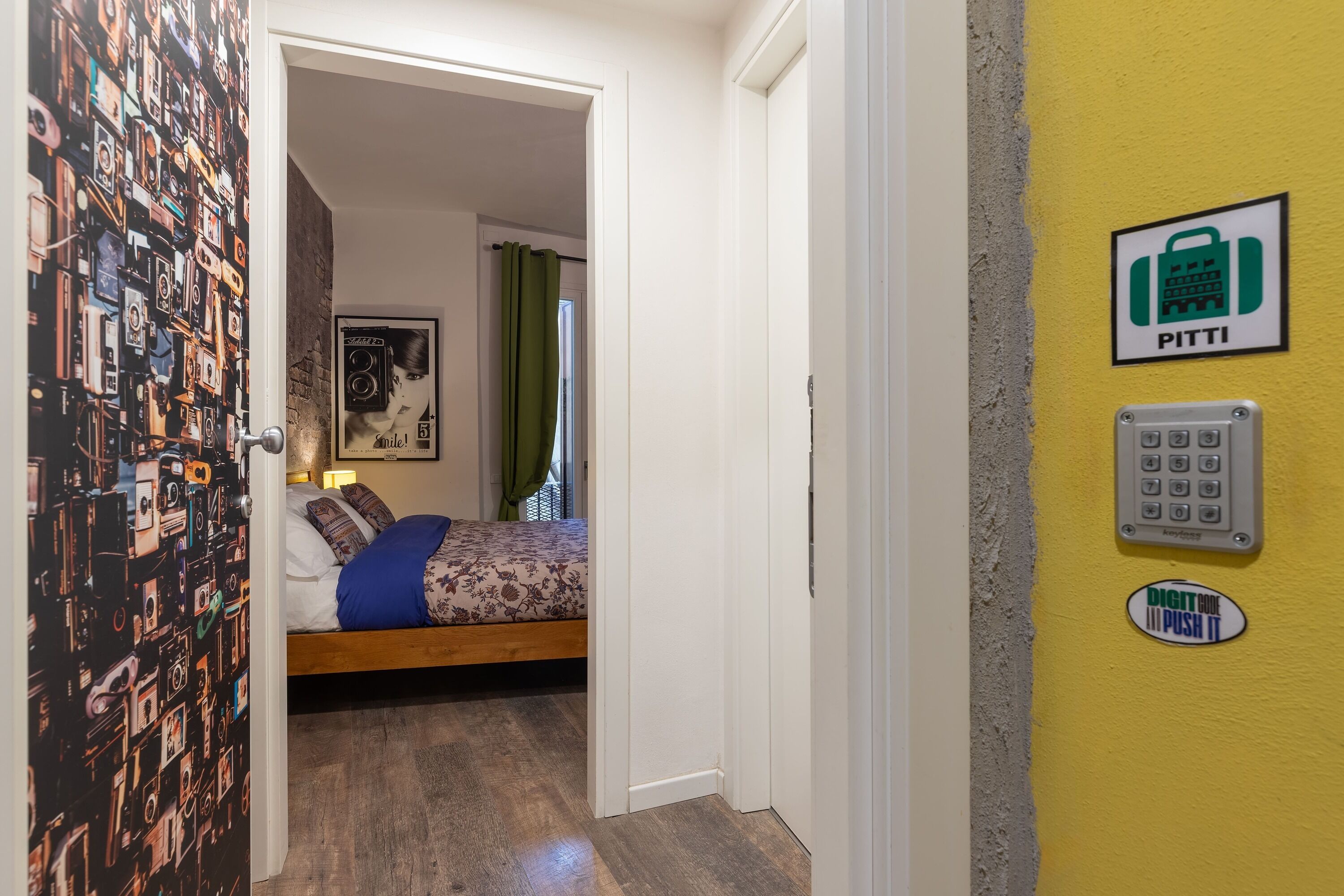 Ett hotellrum med en säng, en dörr med tapet med kameramotiv, en Pitti-skylt och ett digitalt knappsats.