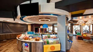 Ein Frühstücksbereich mit einer Theke, auf der verschiedene Speisen und Getränke angeboten werden, einem Sitzbereich mit bunten Stühlen und einem modernen Interieur.