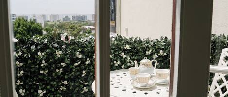 Un balcon donnant sur la ville, une table avec une théière et des tasses, et une chaise en osier blanche.