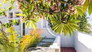 A narrow, white-walled courtyard with a red-tiled floor, surrounded by lush green foliage and flowering plants. - Vesper Apartments (Cape Town)
