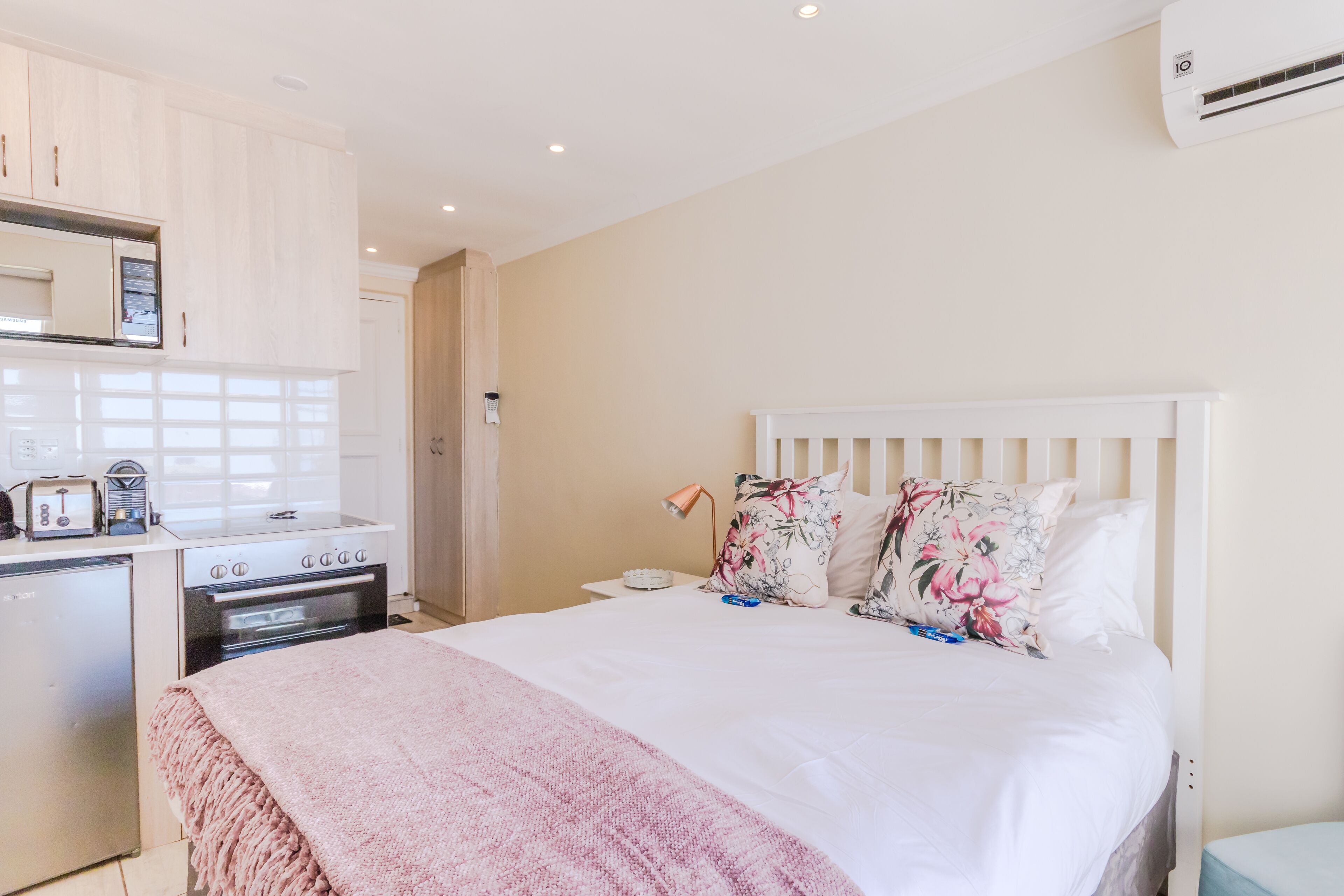 A bedroom with a bed, bedside table, lamp, and a kitchenette area with a stove and microwave.
