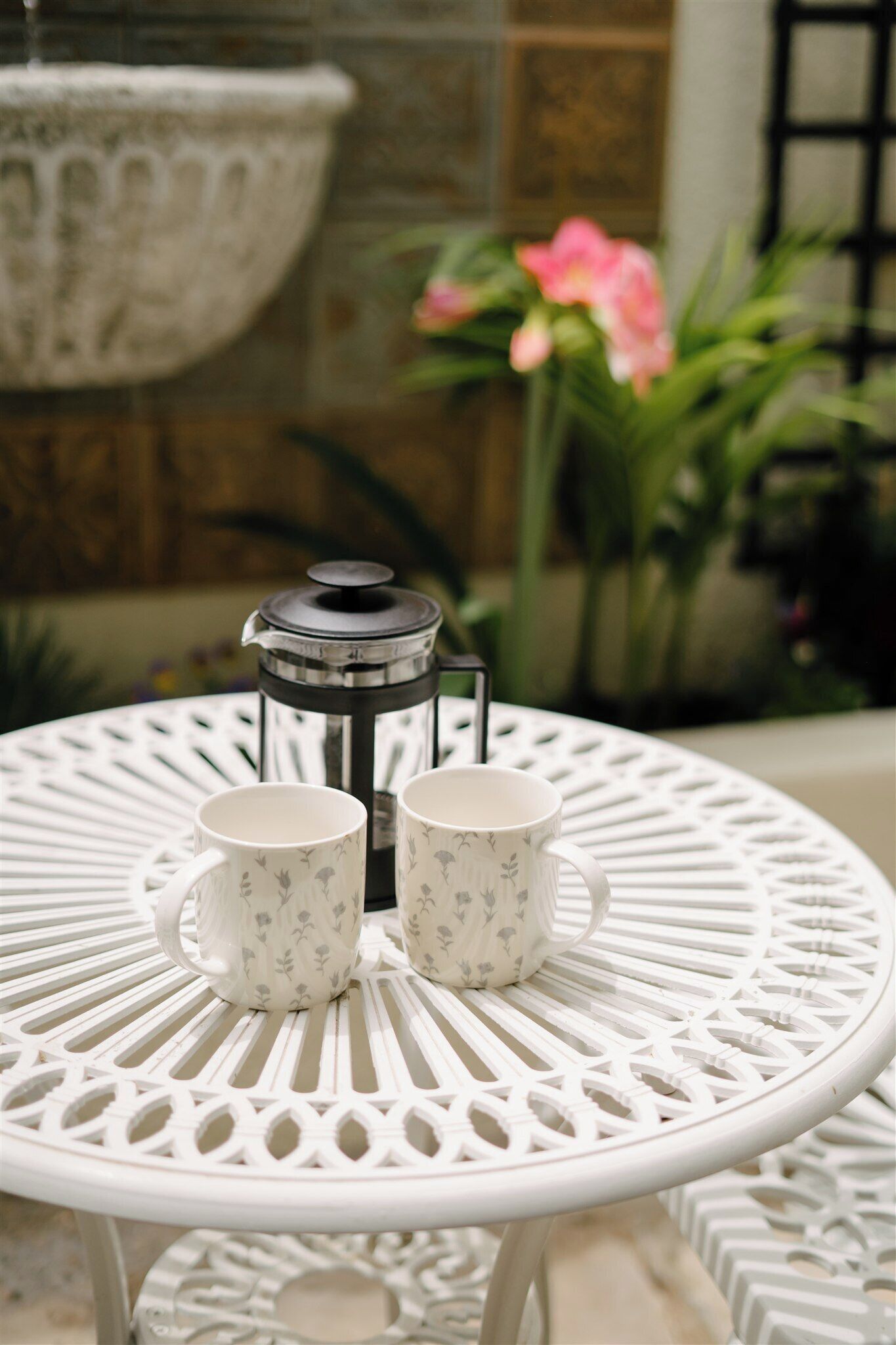 A white metal table with a decorative pattern, two white mugs with floral designs, and a metal lantern.